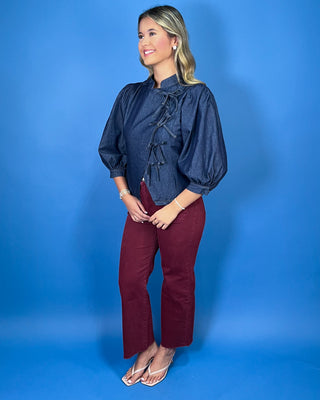 Dark wash denim tie-front top featuring a structured stand collar, puff three-quarter sleeves, asymmetrical front, and statement bow-tie closures.