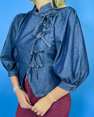 Dark wash denim tie-front top featuring a structured stand collar, puff three-quarter sleeves, asymmetrical front, and statement bow-tie closures.