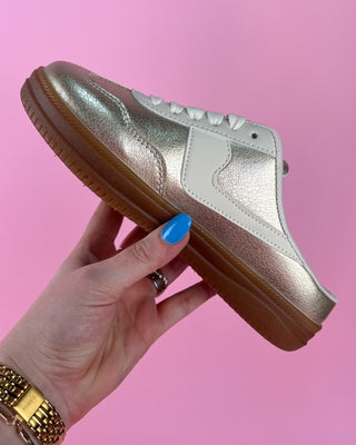 Gold sneaker held by a hand with blue nail polish against a pink background
