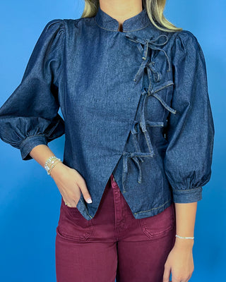 Dark wash denim tie-front top featuring a structured stand collar, puff three-quarter sleeves, asymmetrical front, and statement bow-tie closures.