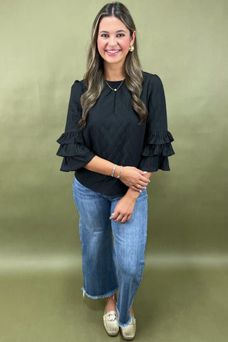 Woman wearing a black ruffled blouse and blue jeans against a green background