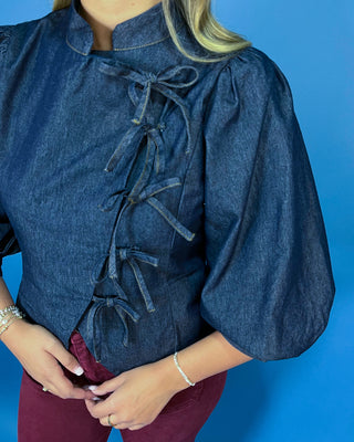 Dark wash denim tie-front top featuring a structured stand collar, puff three-quarter sleeves, asymmetrical front, and statement bow-tie closures.