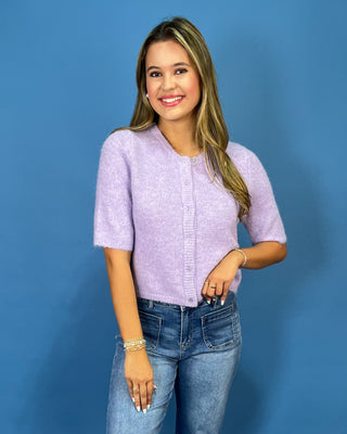 A woman modeling a short-sleeve lavender knit cardigan with a button-up front, paired with blue jeans, against a solid blue studio background.