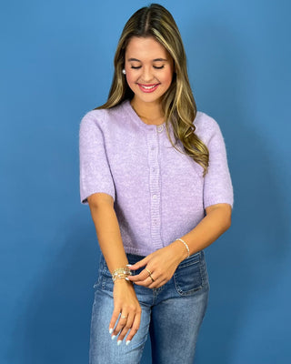 A woman wearing a short-sleeve lavender knit cardigan and blue jeans, standing against a solid blue background.