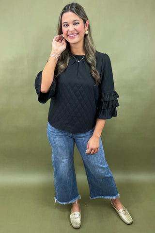 Woman wearing a black top with ruffled sleeves and blue jeans on a green background