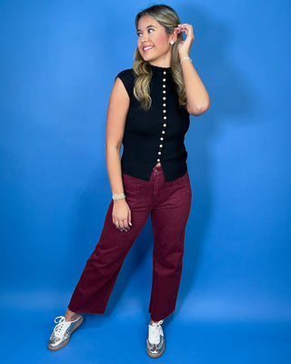 Black ribbed knit sleeveless top featuring a mock neckline, contrasting button-front detail, fitted silhouette, and pointed split hem.