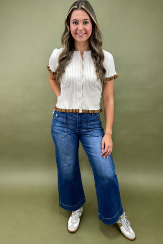 Woman wearing a white blouse with ruffled sleeves, blue jeans, and white sneakers on a green background