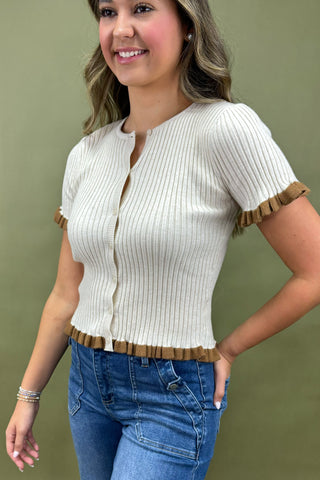 Woman wearing a white ribbed sweater with brown trim and blue jeans against a green background