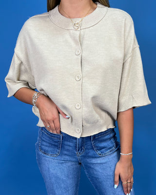 A woman wearing a cream-colored knit cardigan with short sleeves and front buttons, paired with blue jeans, set against a solid blue background.