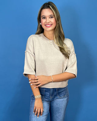 A woman modeling a beige knit sweater with elbow-length sleeves and a crew neck, paired with blue jeans, set against a solid blue studio background.
