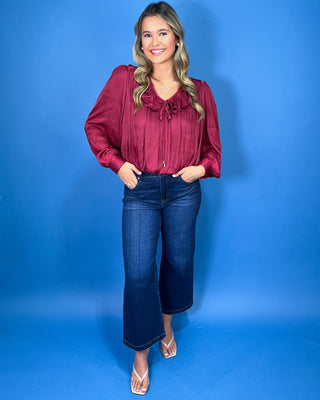 Burgundy satin blouse featuring a romantic ruffle neckline, front tie detail, long balloon sleeves, and a softly gathered hem.