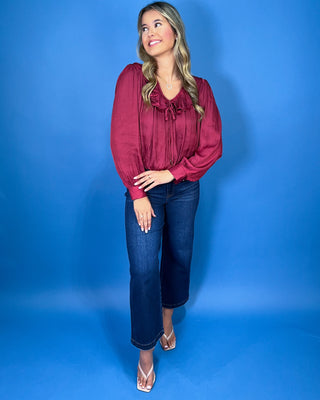 Burgundy satin blouse featuring a romantic ruffle neckline, front tie detail, long balloon sleeves, and a softly gathered hem.