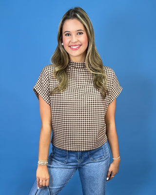 A woman modeling a brown and beige gingham short-sleeve mock neck top with blue jeans, standing against a solid blue background.