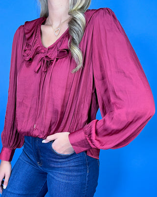 Burgundy satin blouse featuring a romantic ruffle neckline, front tie detail, long balloon sleeves, and a softly gathered hem.