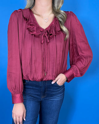 Burgundy satin blouse featuring a romantic ruffle neckline, front tie detail, long balloon sleeves, and a softly gathered hem.