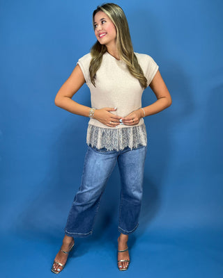 A woman modeling a cream-colored knit top with a lace hem, paired with blue jeans and metallic sandals, standing against a solid blue studio backdrop.