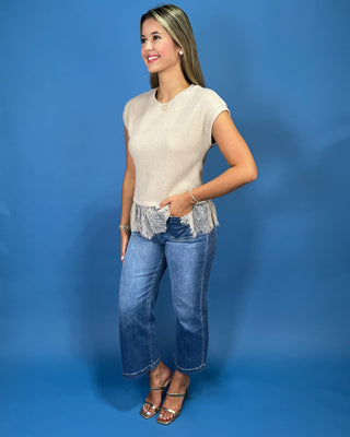 A woman modeling a cream-colored short-sleeve knit top with a frayed lace hem, paired with blue jeans and metallic sandals, set against a solid blue studio backdrop.