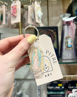 Hand holding a keychain with a tag displaying 'Pitre Art' in a store setting.