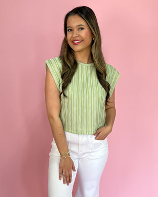 Woman wearing a green patterned top and white pants against a pink background