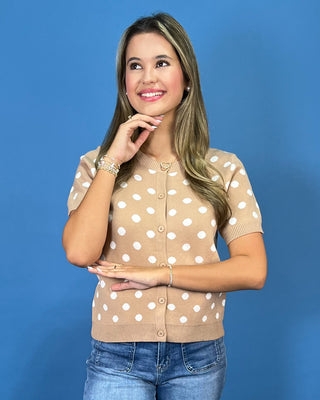 A woman modeling a beige short-sleeve cardigan sweater with white polka dots, paired with blue jeans, set against a solid blue studio background.