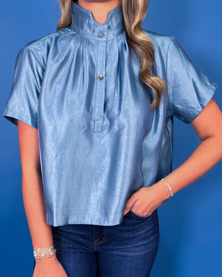 Light blue metallic blouse featuring a structured stand collar, pleated neckline, gold button accents, short sleeves, and a relaxed silhouette.