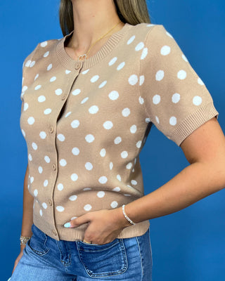 A woman wearing a beige short-sleeve cardigan with white polka dots, paired with blue jeans, set against a solid blue background.