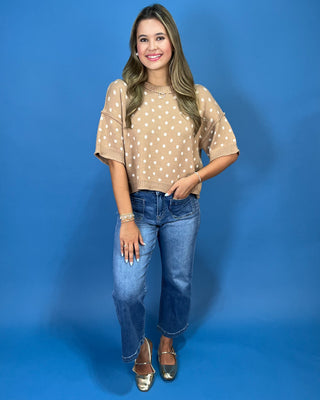 A woman modeling a beige short-sleeve knit top with white polka dots, paired with blue jeans and metallic gold flats, standing against a solid blue studio background.