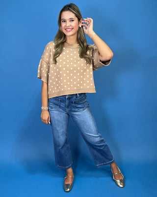 A woman modeling a beige short-sleeve knit top with white polka dots, paired with blue jeans and metallic silver flats, standing against a solid blue studio backdrop.