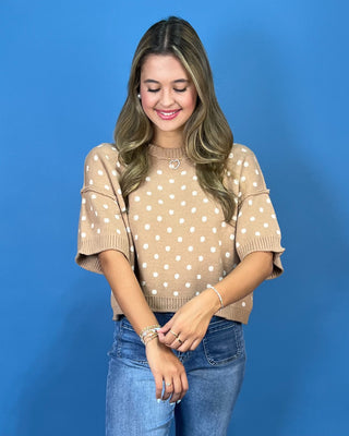 A woman wearing a beige short-sleeve knit sweater with white polka dots and blue jeans, standing against a solid blue background.