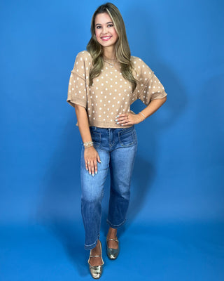 A woman modeling a beige short-sleeve top with white polka dots, paired with blue jeans and metallic shoes, standing against a solid blue studio background.