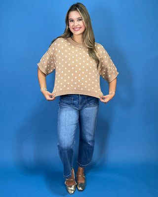 A woman modeling a beige short-sleeve knit sweater with white polka dots, paired with blue jeans and metallic gold flats, standing against a solid blue studio backdrop.