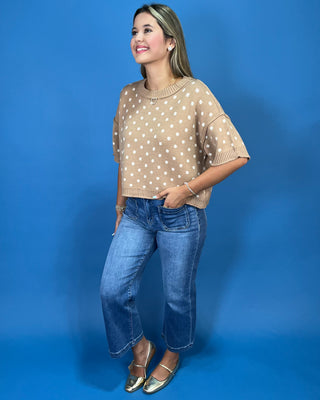 A woman modeling a beige short-sleeve knit sweater with white polka dots, paired with blue jeans and metallic gold flats, standing against a solid blue studio backdrop.