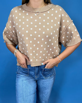 A woman wearing a beige knit sweater with white polka dots and drop shoulders, paired with blue jeans, standing against a solid blue background.