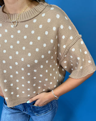 A person wearing a beige knit sweater with white polka dots and blue jeans, standing against a solid blue background.