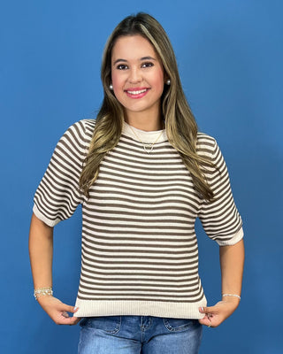 A woman modeling a short-sleeve knit top with horizontal brown and cream stripes, paired with blue jeans, against a solid blue studio background.