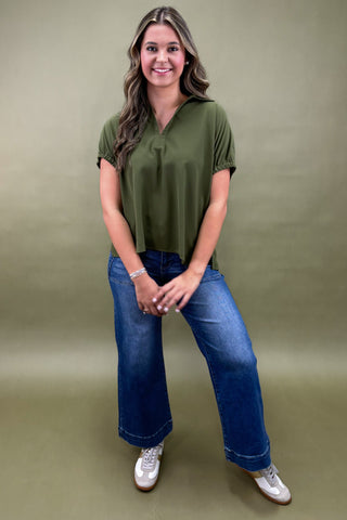 Woman wearing an olive green top and blue jeans against a beige background