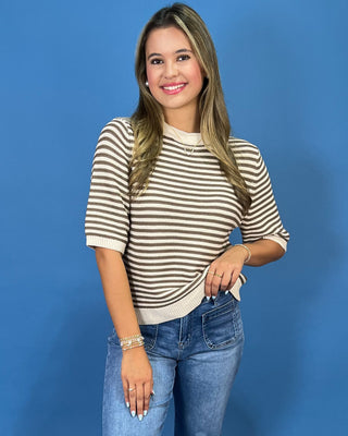 A woman modeling a short-sleeve knit top with horizontal brown and cream stripes, paired with blue jeans, against a solid blue studio background.