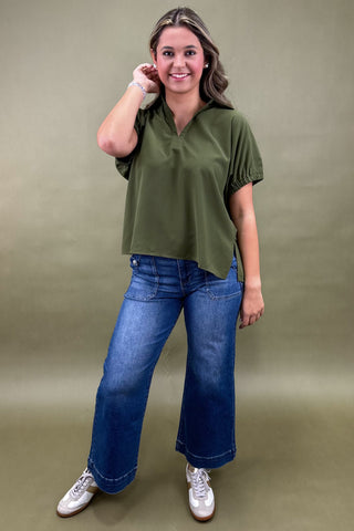 Woman wearing an olive green top and blue jeans against a beige background