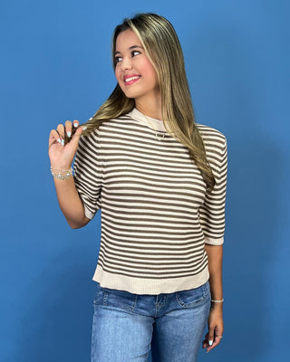 A woman modeling a beige and brown horizontally striped short-sleeve knit top with blue jeans, standing against a solid blue background.
