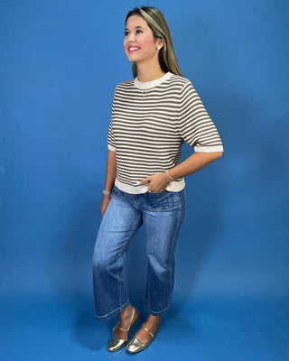 A woman modeling a short-sleeve beige and white striped knit top with blue jeans and metallic flat shoes, standing against a solid blue studio backdrop.