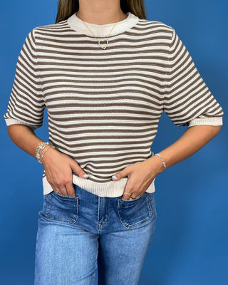 A woman wearing a brown and off-white horizontally striped short-sleeve knit top with a ribbed crew neck, cuffs, and hem, paired with blue jeans, set against a solid blue background.