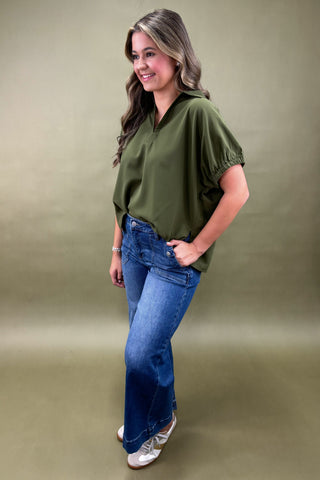 Woman wearing an olive green blouse and blue jeans against a beige background