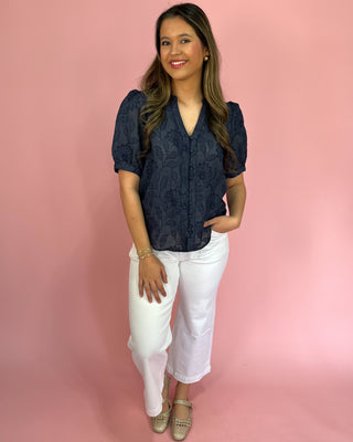 Woman wearing a navy blue lace top and white pants against a pink background
