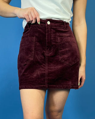 Burgundy corduroy mini skirt featuring a high-rise fit, front patch pockets, button-and-zip closure, and a structured straight silhouette.