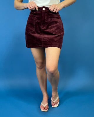 Burgundy corduroy mini skirt featuring a high-rise fit, front patch pockets, button-and-zip closure, and a structured straight silhouette.