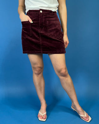 Burgundy corduroy mini skirt featuring a high-rise fit, front patch pockets, button-and-zip closure, and a structured straight silhouette.