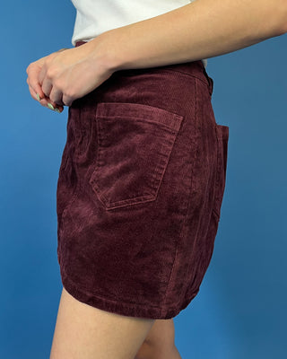 Burgundy corduroy mini skirt featuring a high-rise fit, front patch pockets, button-and-zip closure, and a structured straight silhouette.