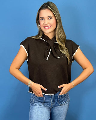 A woman modeling a dark brown short-sleeve knit top with white scalloped trim and a matching bow tie neckline, paired with blue jeans against a solid blue background.