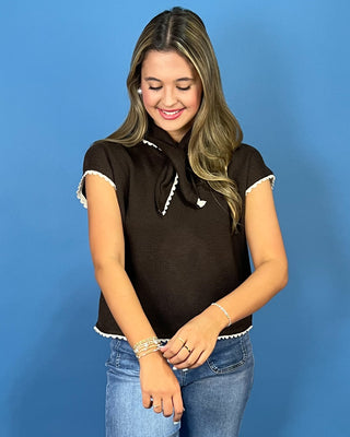 A woman wearing a dark brown short-sleeve knit top with white lace trim and a bow detail, paired with blue jeans, standing against a solid blue background.