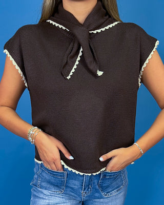 A woman wearing a dark brown ribbed knit top with a wide collar tied in a bow, short sleeves with white scalloped trim, and a white scalloped hem, paired with blue jeans against a solid blue background.